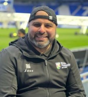 Photo of Anwar. Anwar is sat in a football stadium, he is facing the camera and is smiling. Anwar has dark skin and a grey beard. He is wearing a grey tracksuit and a navy blue baseball cap, which he is wearing backwards.