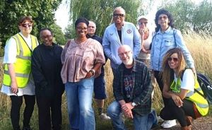 Nine people, male and female, of mixed ages and ethnicity, standing and kneeling facing the camera. The group are outside surrounded by trees and long grass.