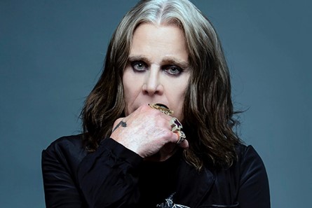 Head and shoulders photo of Ozzy Osbourne looking at camera with his hand on his chin. He is wearing a black top.