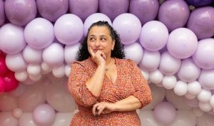 A picture of Nabila posing in front of a colourful balloon display. She has long dark hair and is wearing a light brown dress. She is posing with her hand under her chin and is looking to the left.