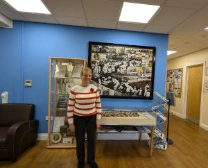 Picture of Linda. Linda has fair skin and short fair hair and is wearing glasses. She is wearing a red and white striped jumper and is stood in front of a bright blue wall and some shelves.