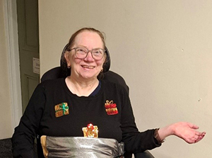 Picture of Lisa. Lisa is wearing a black Christmas jumper with sparkly Christmas presents on it. She is sat down and is smiling at the camera.