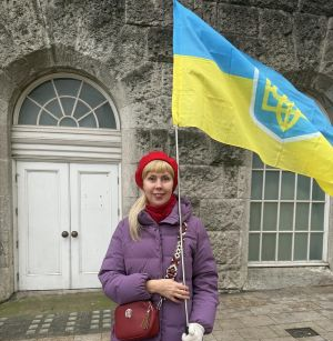Photo of Viktoriia. She is stood outside a stone building holding a large Ukranian flag in her left hand. Viktoriia has shoulder length blonde hair and is wearing a purple coat and a red beret. She is also wearing a red bag across her body.