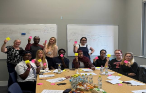 A group of people gathered around a table. Some are standing and some are sat on chairs. The group are diverse in terms of age, ethnicity and gender. They are all holding post-it notes and are looking at the camera. On the table, there bottles of water.